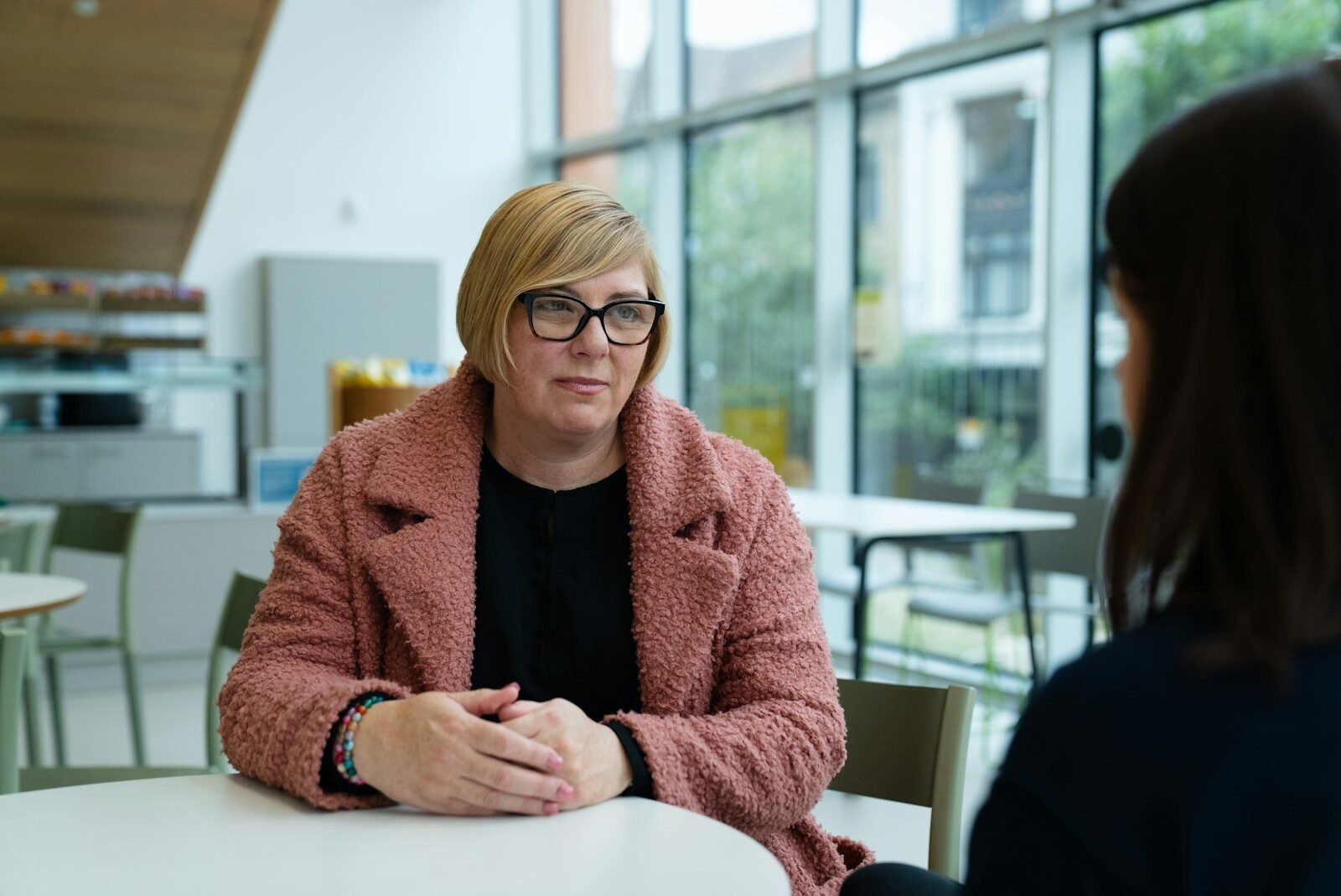 Woman in pink coat talks to someone at table